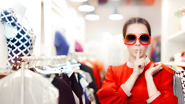 Seorang individu mengenakan pakaian merah mencolok dan kacamata besar berdiri di toko pakaian yang terang, melakukan gestur "diam" dengan jari di bibir, dikelilingi rak baju dan manekin.